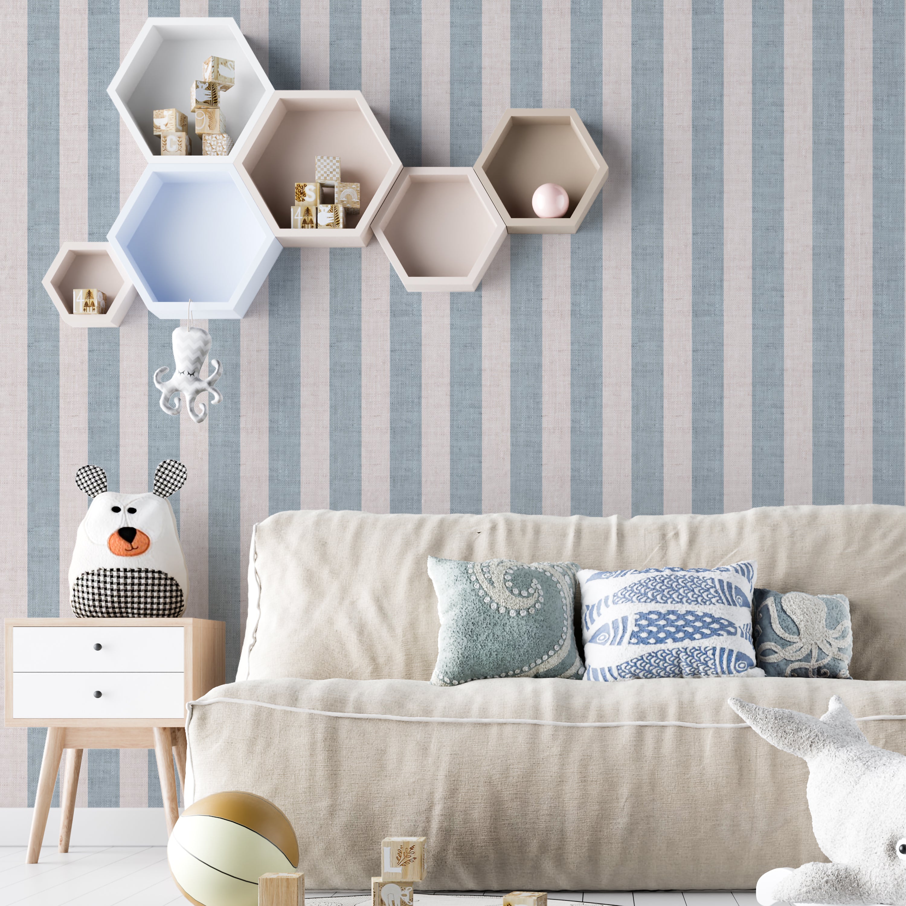 A cozy living room scene showcasing the Ticking Fabrics 7 II Wallpaper with vertical blue and cream stripes. The room includes a neutral-colored sofa with decorative pillows, hexagonal wall shelves, and playful children’s toys, highlighting the wallpaper’s versatility in a domestic setting.