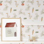 A section of a wall decorated with Farm Friend Wallpaper - Happy Bunnies, featuring whimsical illustrations of bunnies, geese, carrots, radishes, and grass, along with a white shelf and colorful pennant banner.