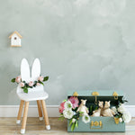 A child's chair with bunny ear backrest, decorated with a garland of pink roses and green leaves, next to an open toy chest filled with plush animals and flowers, against a light sage cloud mural wallpaper.