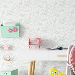 The Watercolor Spring Bird Wallpaper - Seafoam brings a light and airy feel to a child's playroom, its delicate leaf patterns offering a backdrop to a white desk adorned with playful children's accessories in pastel colors.