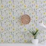 A workspace decorated with the Spring Field Wallpaper - V, showcasing its vibrant pattern of yellow flowers and green foliage. The space includes a wooden wall clock and a white desk lamp beside a small plant.