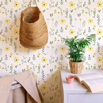 A cozy room featuring the Spring Field Wallpaper - V with a soft white background adorned with bright yellow flowers and green leaves, complemented by a woven basket hanging on the wall and a small potted plant on a white desk