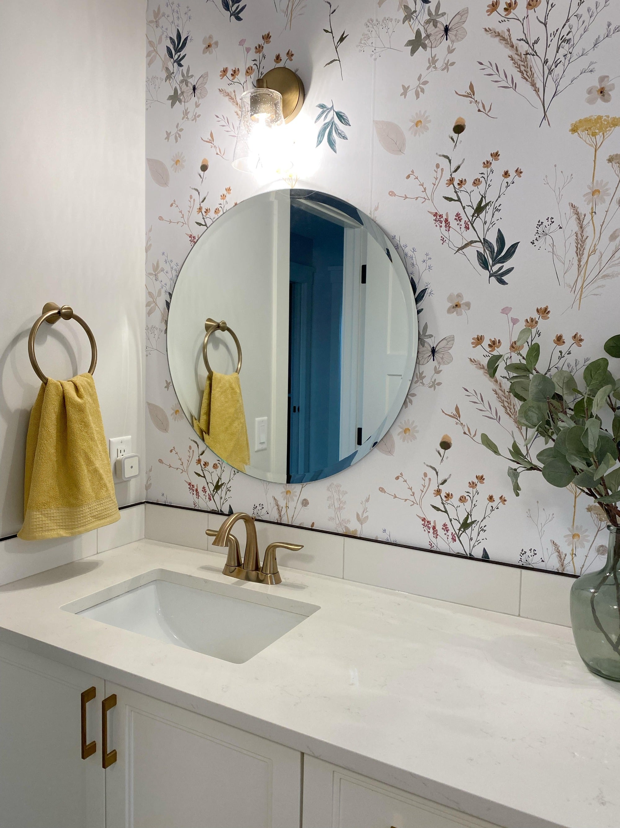 A bright bathroom interior with botanical-themed wallpaper showcasing wildflowers and foliage in muted colors. The space includes a white vanity with dual sinks, gold fixtures, round mirrors, and decorative touches such as a 'brush your teeth' sign and yellow towels.