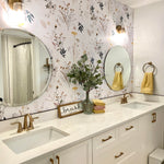 A bright bathroom interior with botanical-themed wallpaper showcasing wildflowers and foliage in muted colors. The space includes a white vanity with dual sinks, gold fixtures, round mirrors, and decorative touches such as a 'brush your teeth' sign and yellow towels.