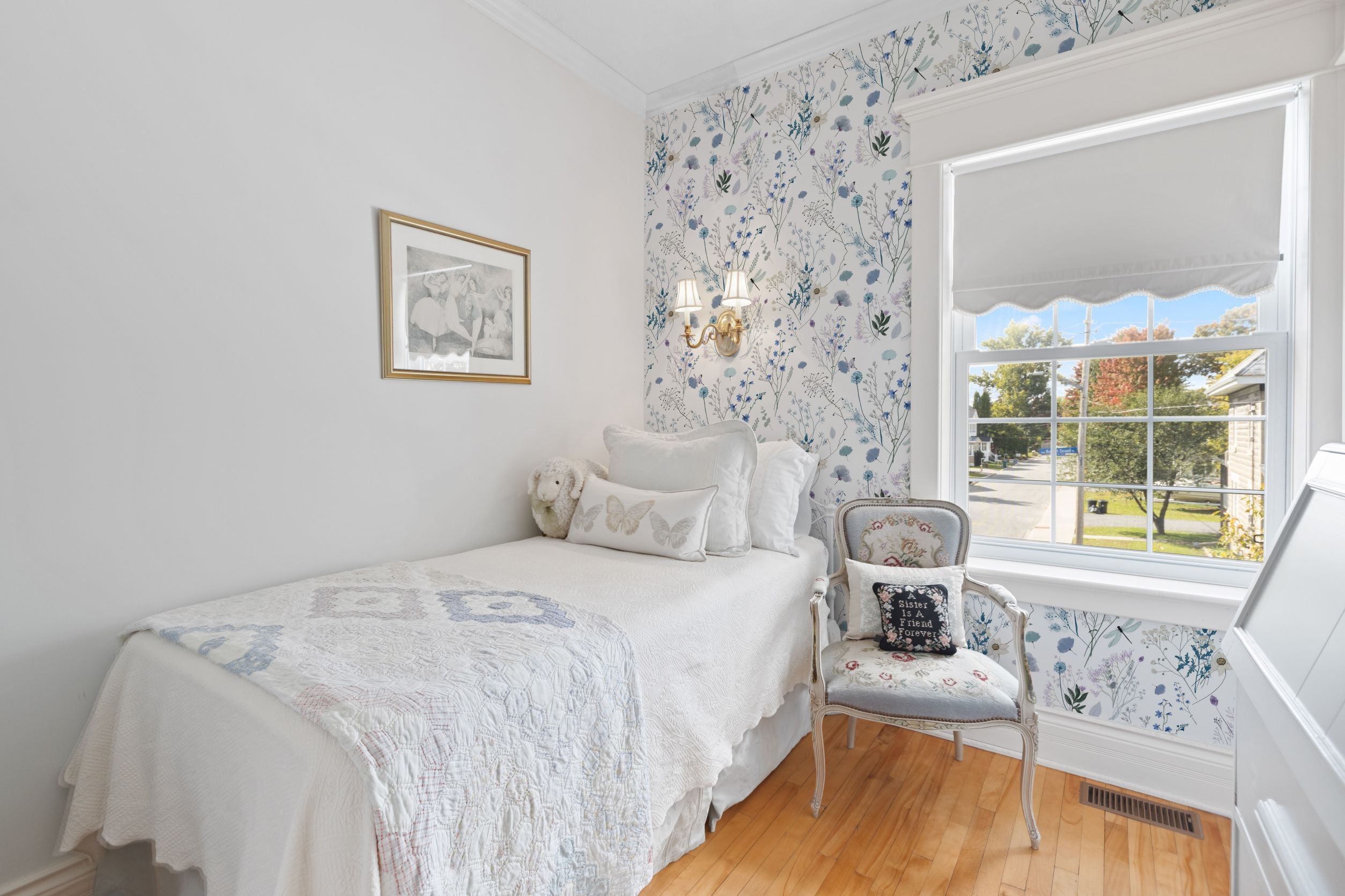 A charming vintage bedroom with natural light streaming through the window, showcasing the Aerie Floral II Wallpaper with delicate blue and purple flowers and soft greenery, complementing a classic white bedspread and antique-style armchair.