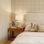 A serene bedroom featuring a headboard upholstered in a light textured fabric. The wall behind is adorned with a gingham patterned wallpaper in shades of beige and white, complemented by a wooden nightstand and a ceramic table lamp. The bed is dressed in soft linen with neutral-toned pillows.