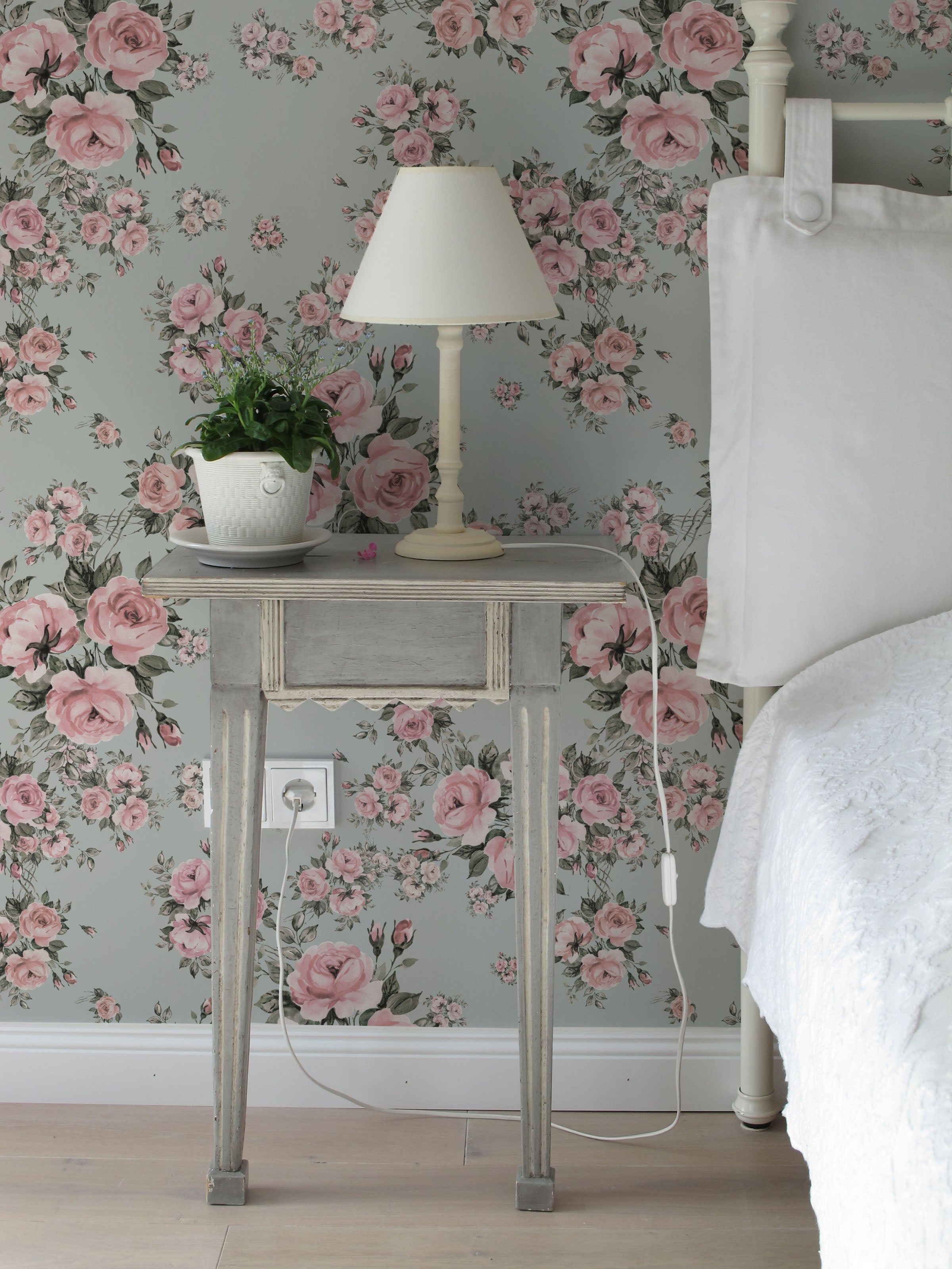 A charming bedroom with Rose Bouquet Wallpaper II featuring pink roses and green leaves on a light blue background. The decor includes a vintage nightstand, a white lamp, and a potted plant