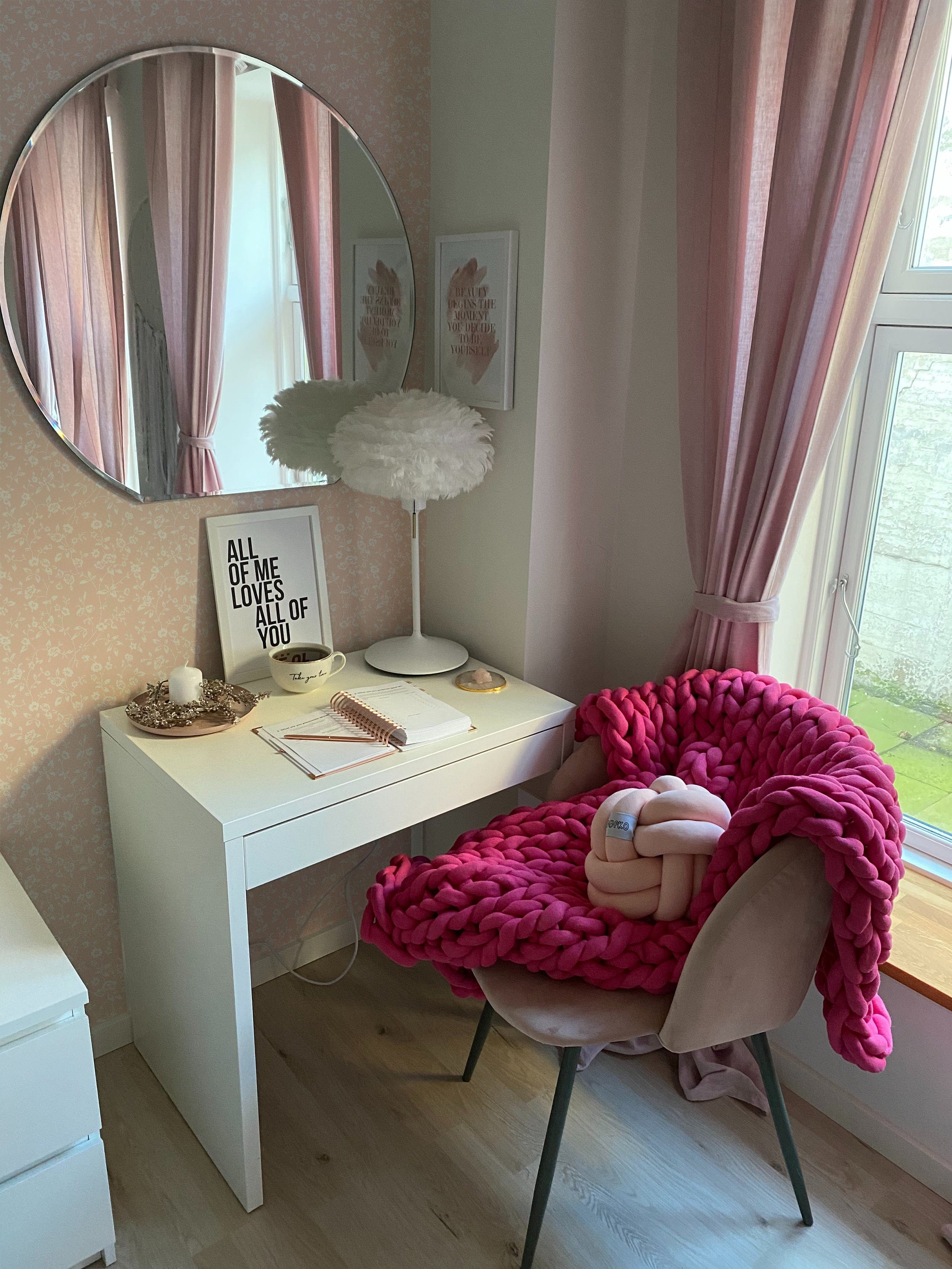 A cozy nook with a white desk and a chair draped with a chunky pink blanket. The wall is adorned with Simple Mauve Floral Wallpaper, complementing the pink curtains and creating a warm, feminine atmosphere. A round mirror and inspirational quotes complete the look.