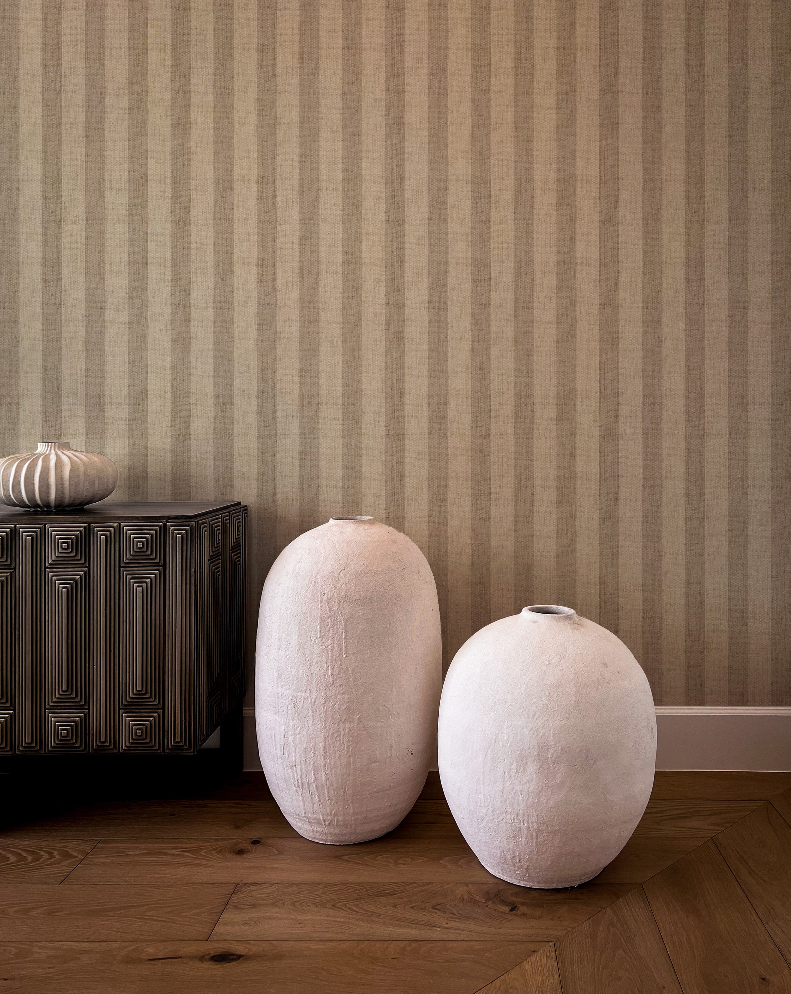 Modern interior featuring Classic Striped Wallpaper in beige tones, complemented by artistic ceramic vases on a hardwood floor. The room's aesthetic is enriched with a dark wooden cabinet and a textured silver vase, creating a sophisticated and contemporary look.