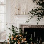 Chic traditional fireplace adorned with beige dainty floral wallpaper, elegantly contrasted by lush greenery and an arrangement of candles and flowers, creating a romantic ambiance