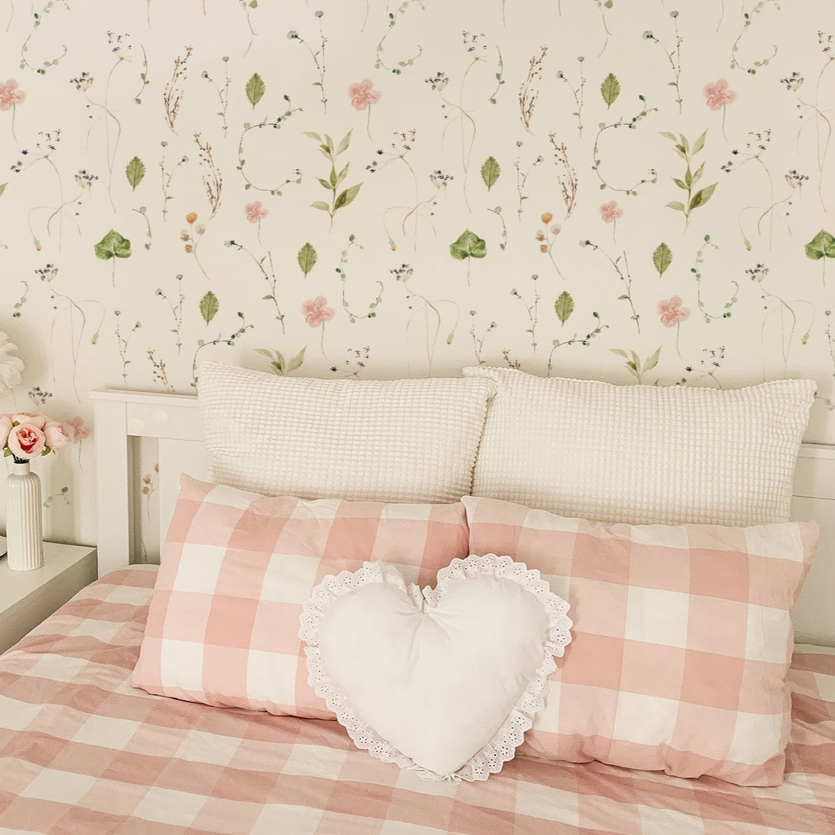 A cozy and romantic bedroom scene highlighted by the Modern Watercolour Floral Wallpaper in Dusty Pink on the walls. The wallpaper's soft pastel flowers and leaves create a gentle and tranquil ambiance. The room is styled with a checkered pink and white bedding, decorative pillows, and fresh flowers, emphasizing a feminine and dreamy interior design.