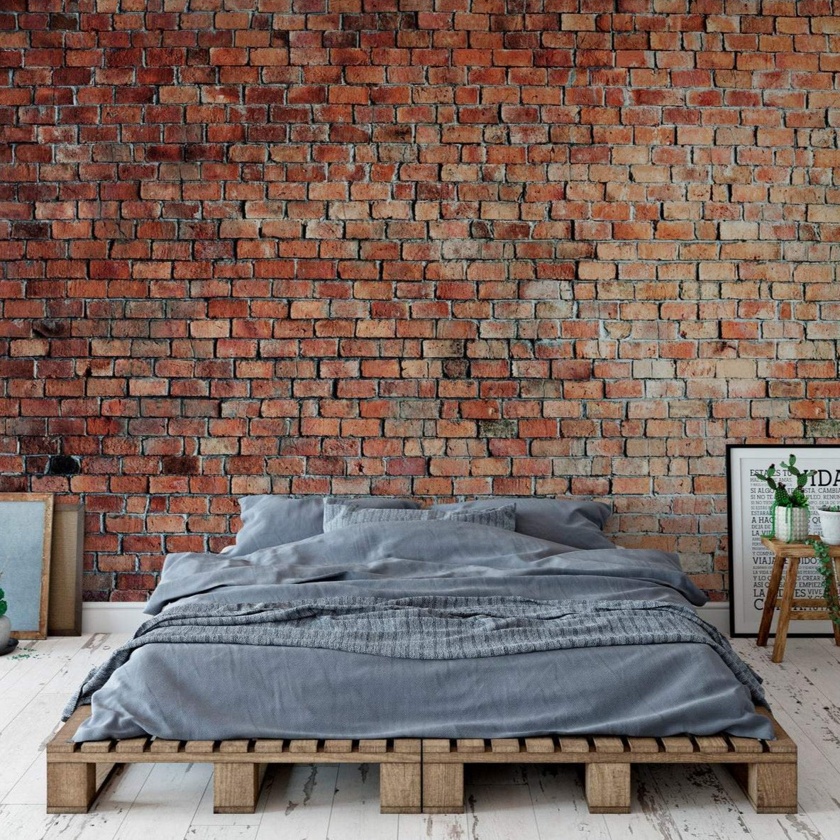 A stylish bedroom setting with Realistic Red Brick Wallpaper as a statement wall behind a bed with grey linen and a wooden pallet base. The brick wall adds a cozy and industrial charm to the room.