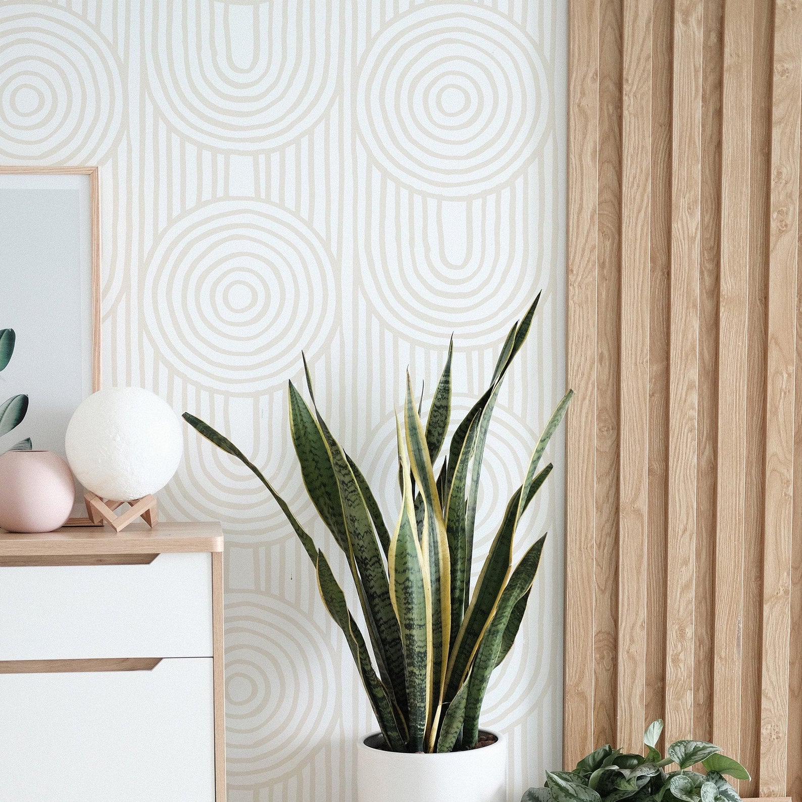 A modern and serene interior featuring the Zen Abstract Wallpaper, with a pattern of neutral-toned concentric circles and elongated ovals on a light background. The design adds a subtle organic touch to the room, complemented by wooden furniture and green houseplants.