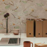 A detailed workspace setting showing a laptop on a wooden desk against a backdrop of soft beige wallpaper adorned with delicate watercolor magnolia flowers, creating a peaceful working environment.