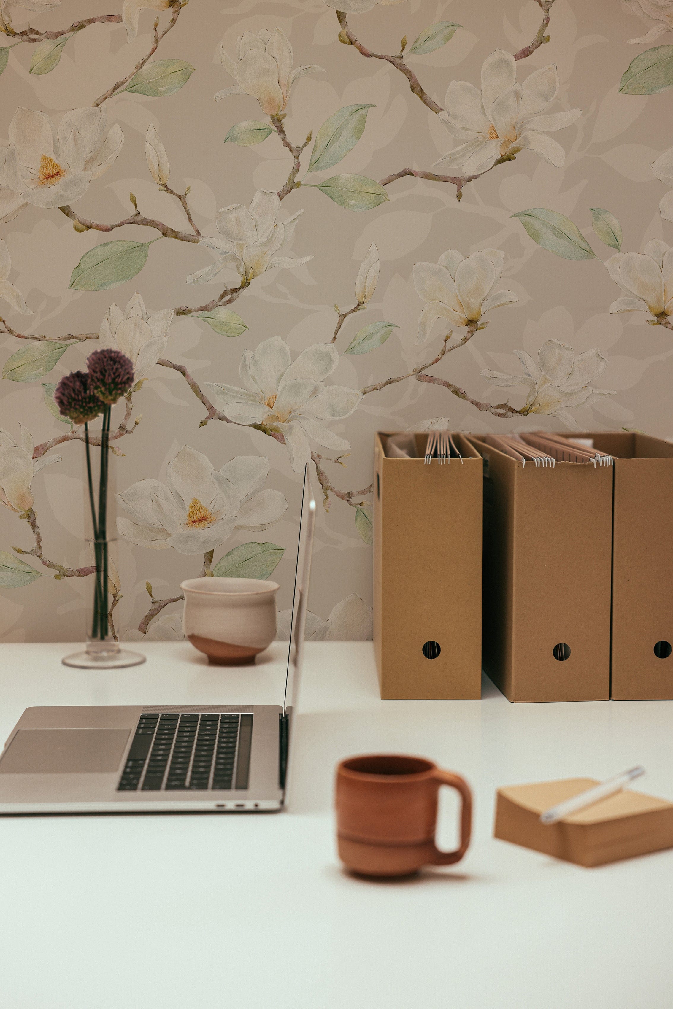 A detailed workspace setting showing a laptop on a wooden desk against a backdrop of soft beige wallpaper adorned with delicate watercolor magnolia flowers, creating a peaceful working environment.