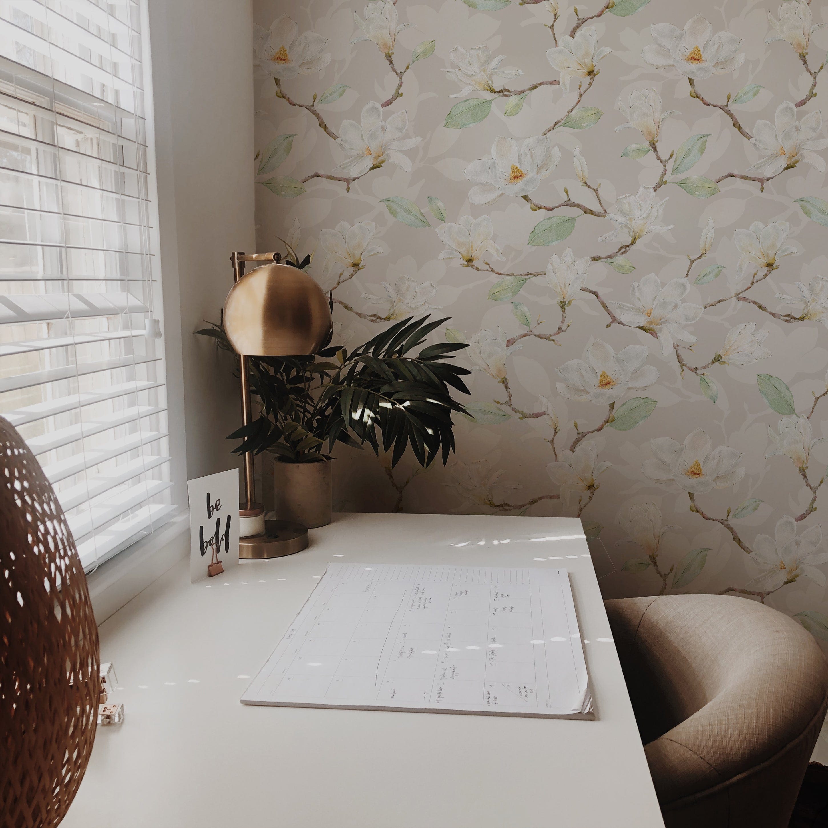 A tranquil corner of a room illuminated by natural light from a window, showcasing an elegant floral wallpaper with large magnolia flowers in soft hues, complemented by a modern desk lamp and indoor plants.