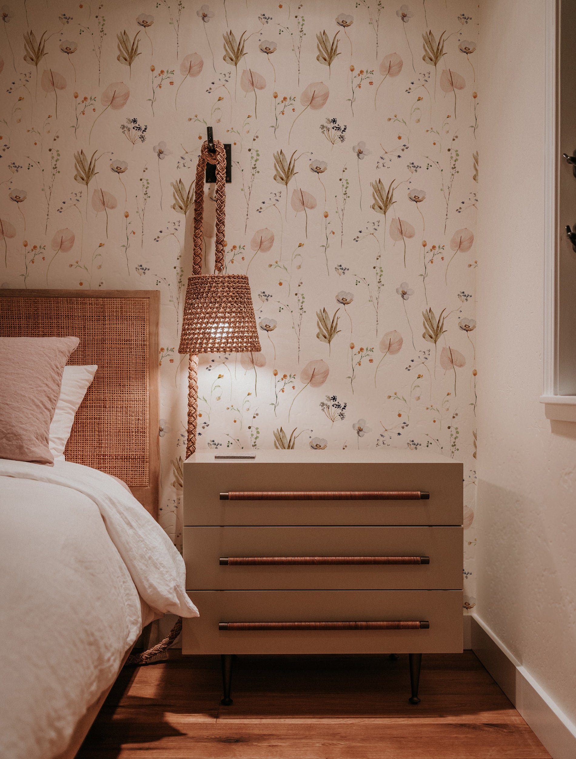 A warmly lit bedroom corner with Ikebana Floral Wallpaper adding a touch of nature-inspired elegance through its delicate floral design, paired with a wicker headboard and a modern nightstand with copper handles.