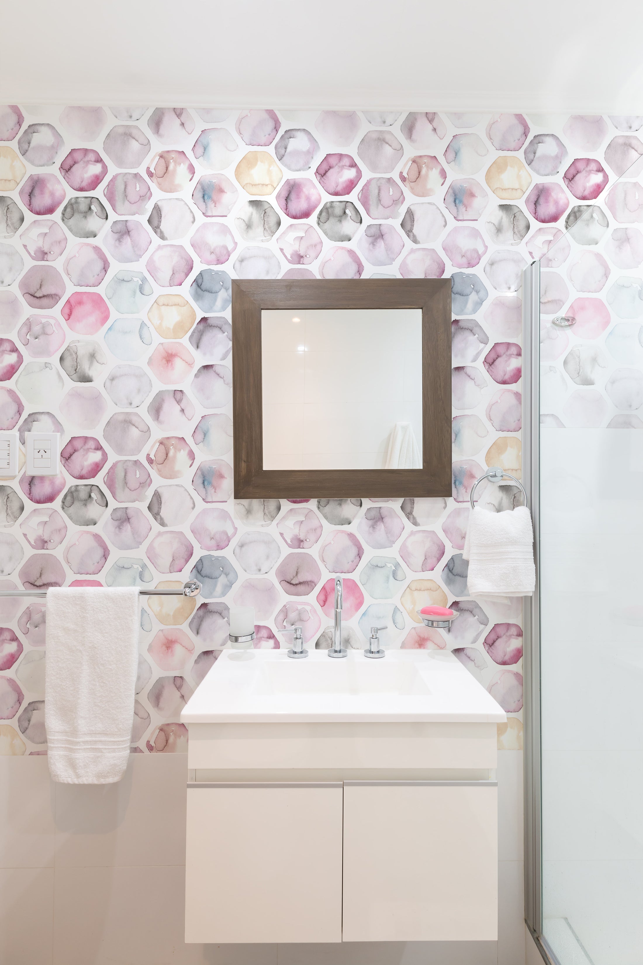 A modern bathroom interior showcasing the Watercolour Honeycomb Wallpaper with a unique pattern of soft watercolor shapes resembling a honeycomb, in muted shades of pink, purple, blue, and beige, adding an artistic and soothing ambiance.