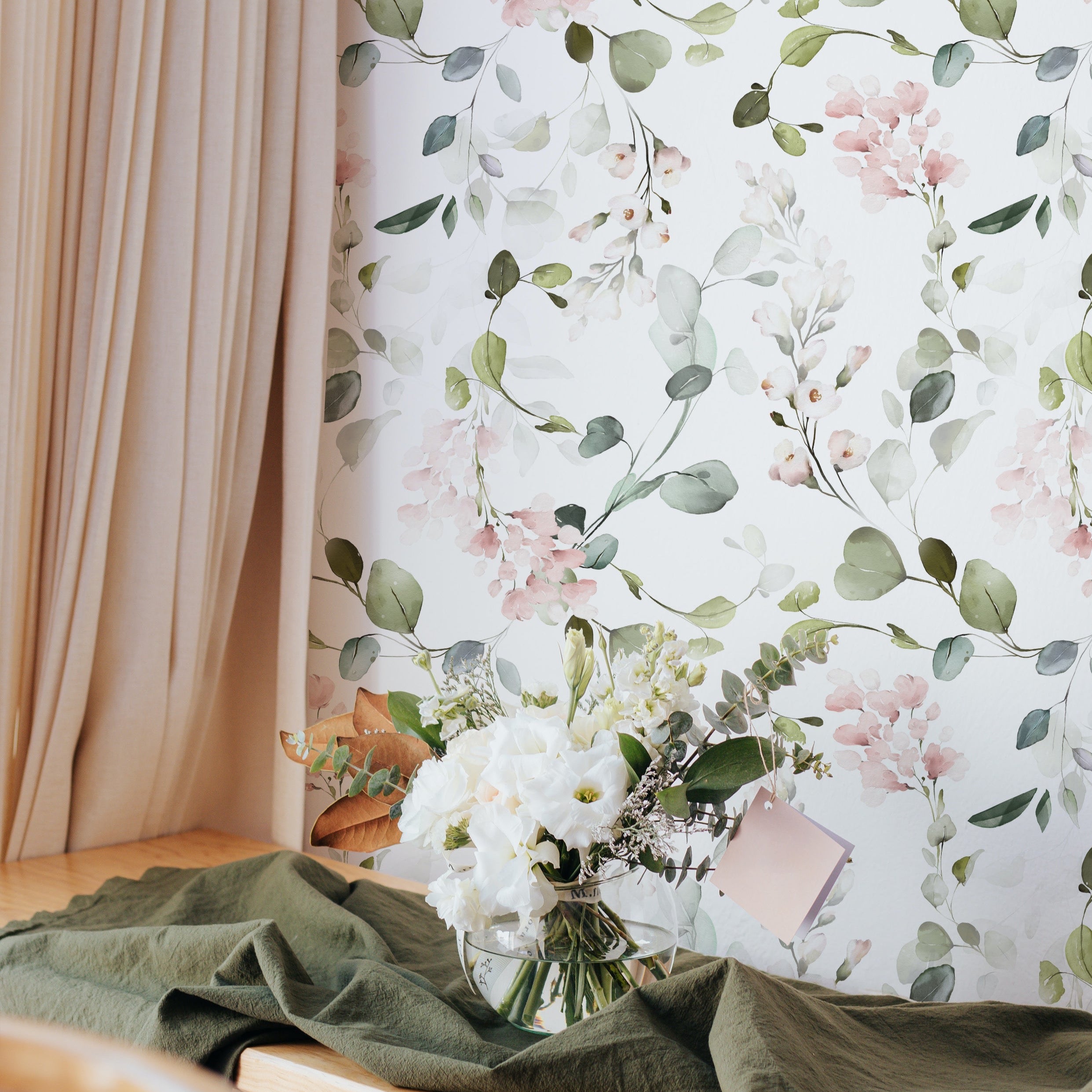 A corner of a bright room showcasing the Pink Floral & Herbs Wallpaper, paired with a wooden table dressed with a sage green throw and a bouquet of white flowers. The graceful floral pattern exudes a calm and sophisticated atmosphere.