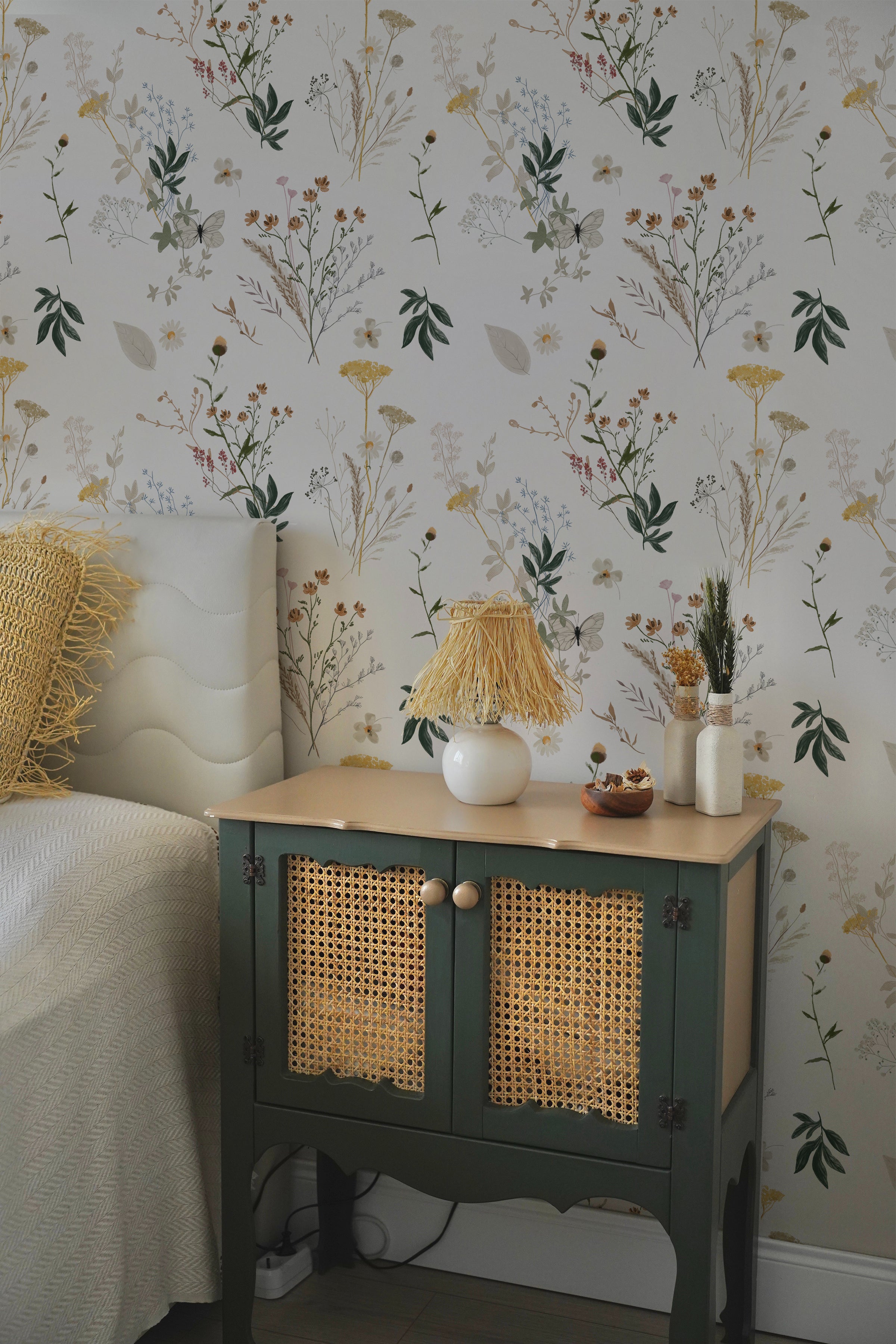 A cozy bedroom corner with a botanical wallpaper featuring a variety of wildflowers and leaves in a soft color palette. A side table painted in dark green with rattan cabinet doors is adorned with a white vase with straw fringes, a small wooden bowl, and textured vases with dried botanicals.