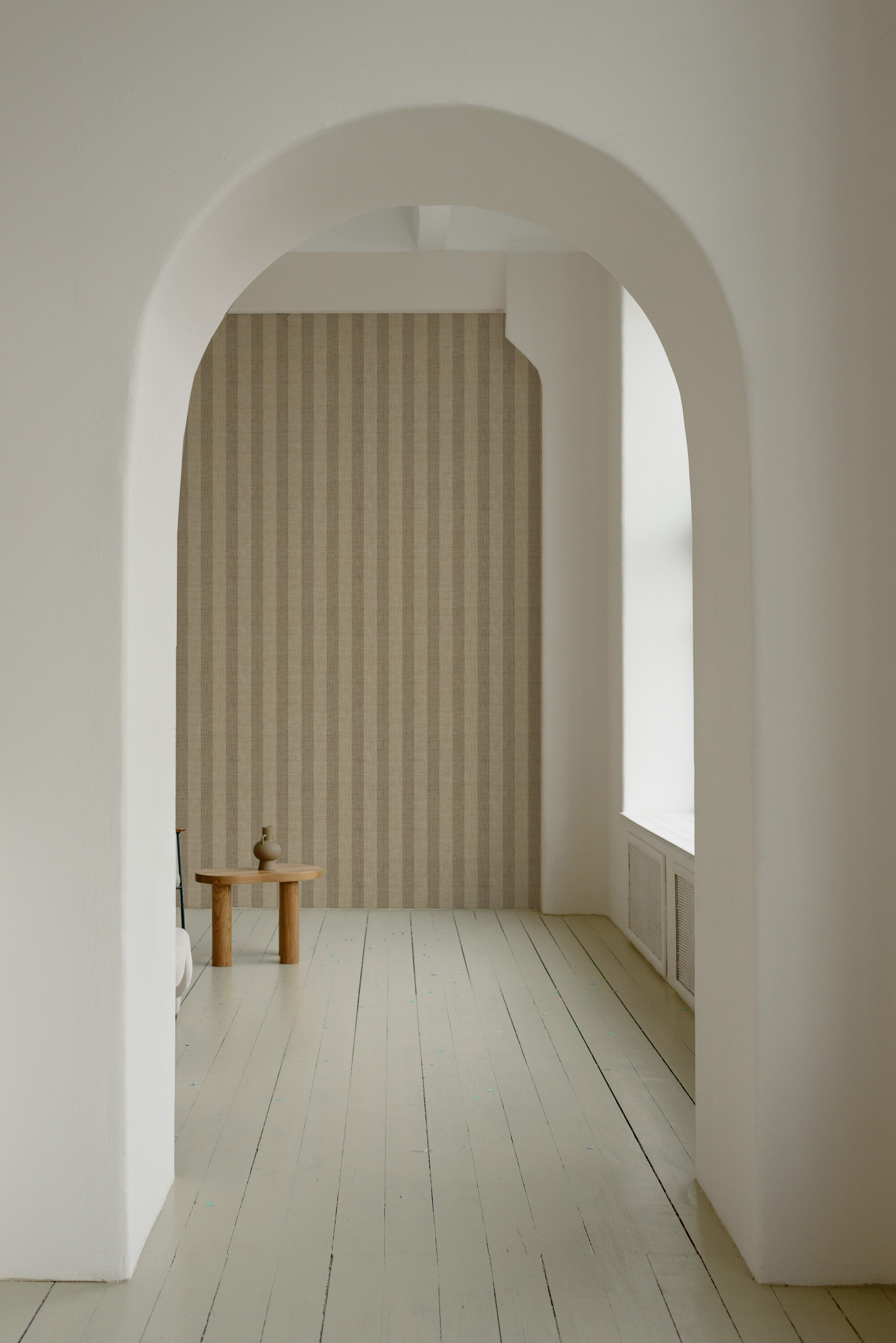 A minimalist archway leads to a room adorned with Classic Striped Wallpaper in neutral beige, enhancing the serene and elegant ambiance. A simple wooden stool and a small ceramic vase sit by the white painted floor, emphasizing the wallpaper's understated elegance.