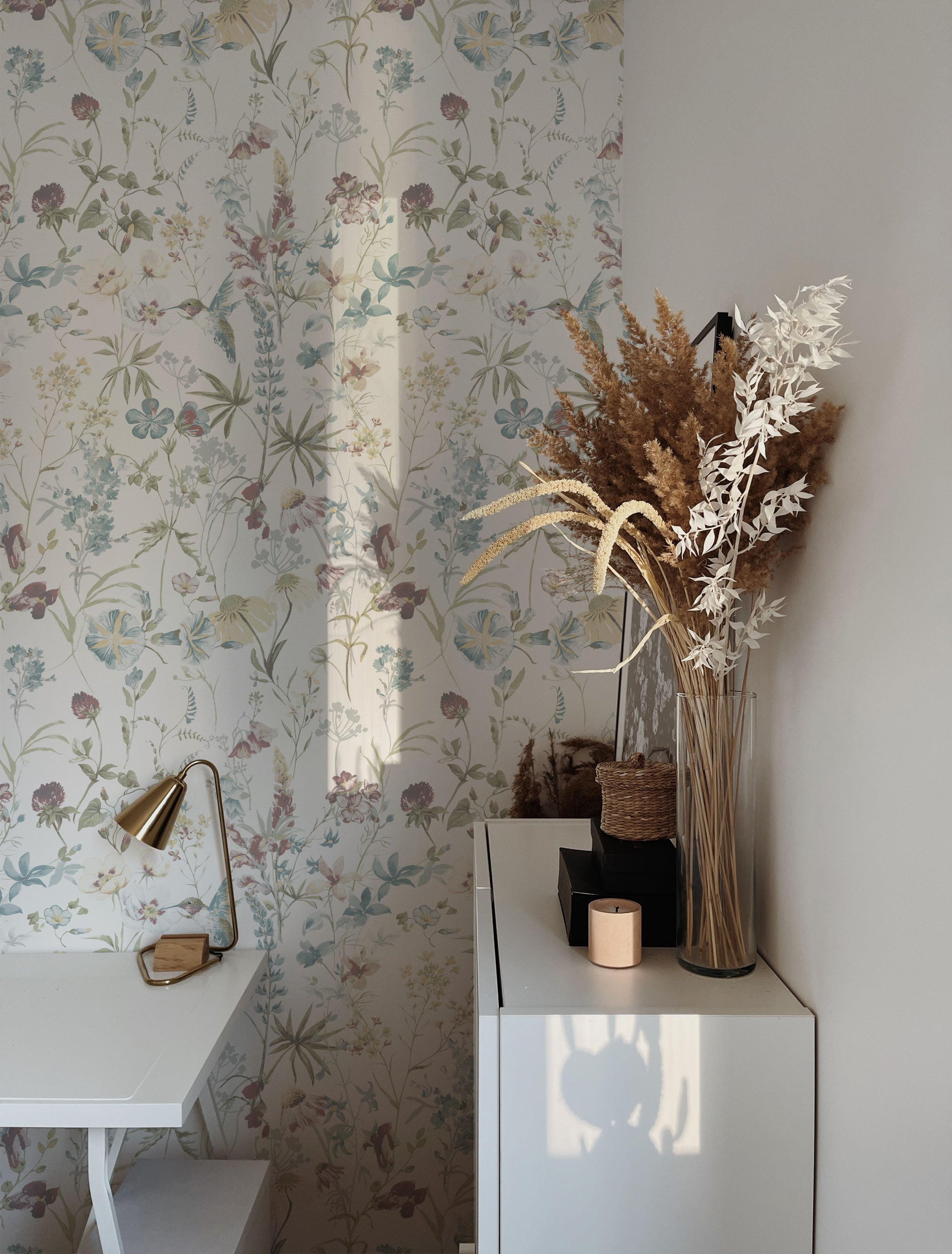 A serene home office corner featuring the Soft Feathered Blooms Wallpaper, which displays a delicate and airy botanical print with flowers and leaves in soft pastel shades of blue, pink, and green on a light background. This wallpaper adds a touch of spring freshness to the room, complemented by natural wood furniture and dried floral arrangements.