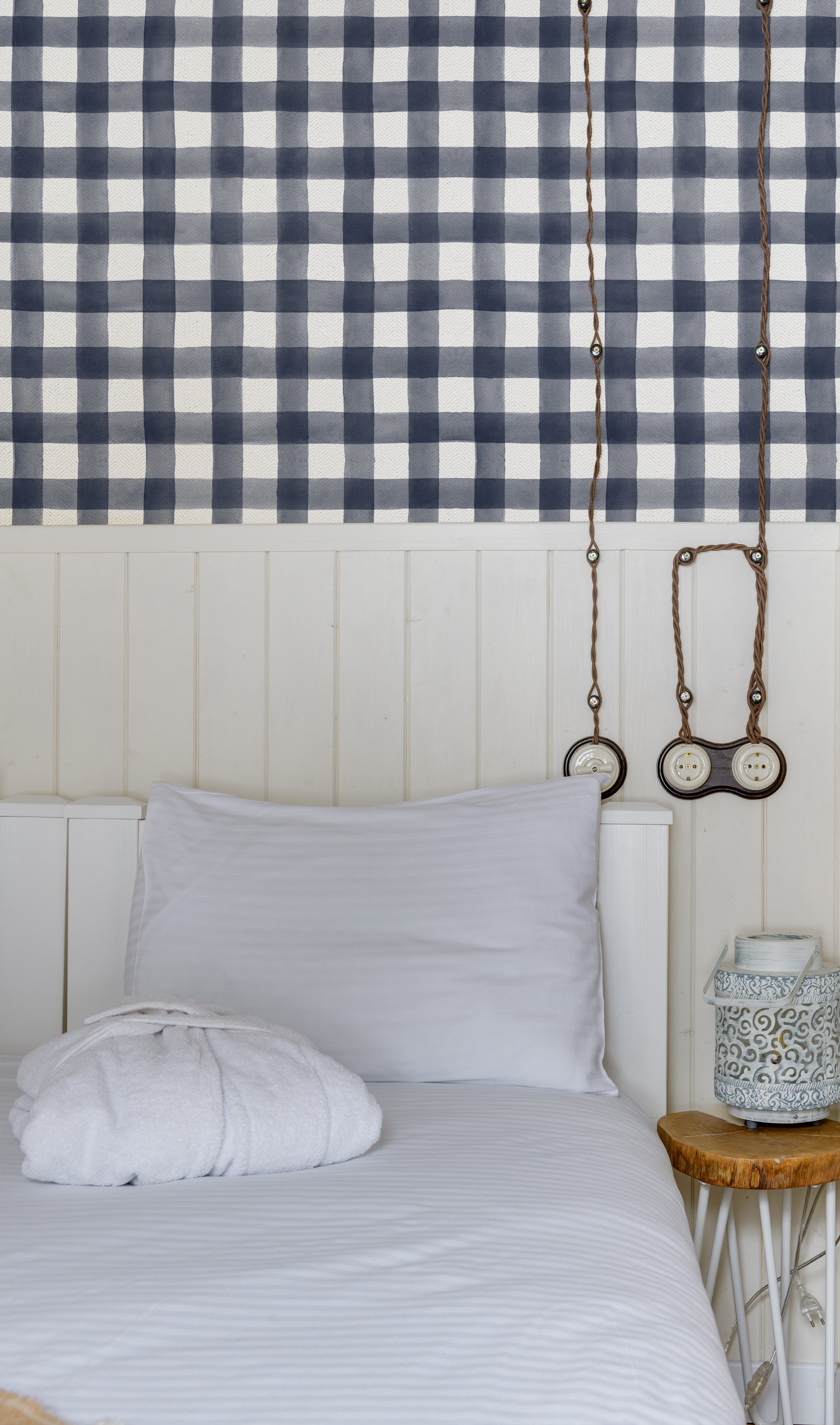 A cozy bedroom setting with a wall covered in navy blue and white buffalo check wallpaper. The room features a single bed with white linens, a white pillow, and a vintage-style round wall clock hanging on the ropes above the bed, creating a classic, inviting look.