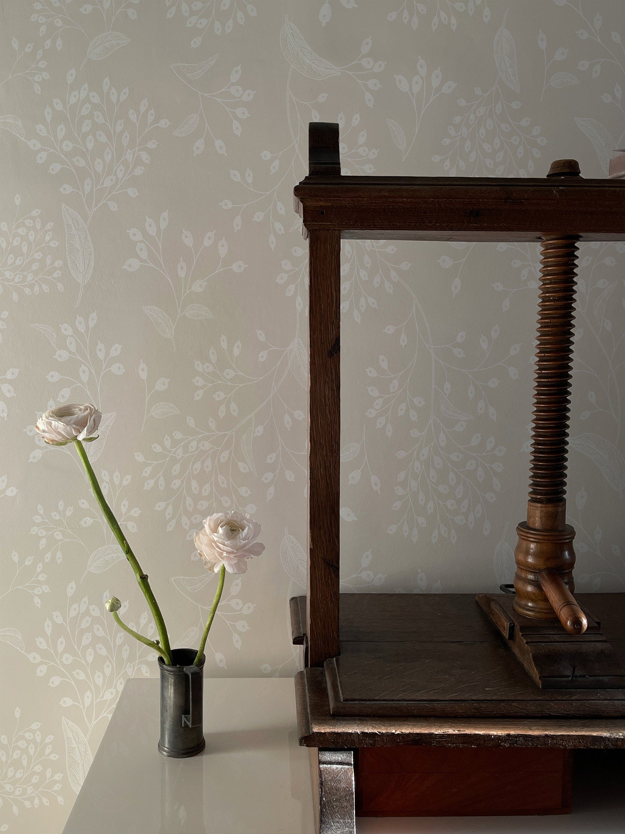A subtle and elegant home decor setup showcasing the Botanical Whisper Wallpaper, which features a delicate pattern of soft beige botanical prints. A dark wooden antique book press and a small vase with two blooming flowers enhance the vintage charm of the room.