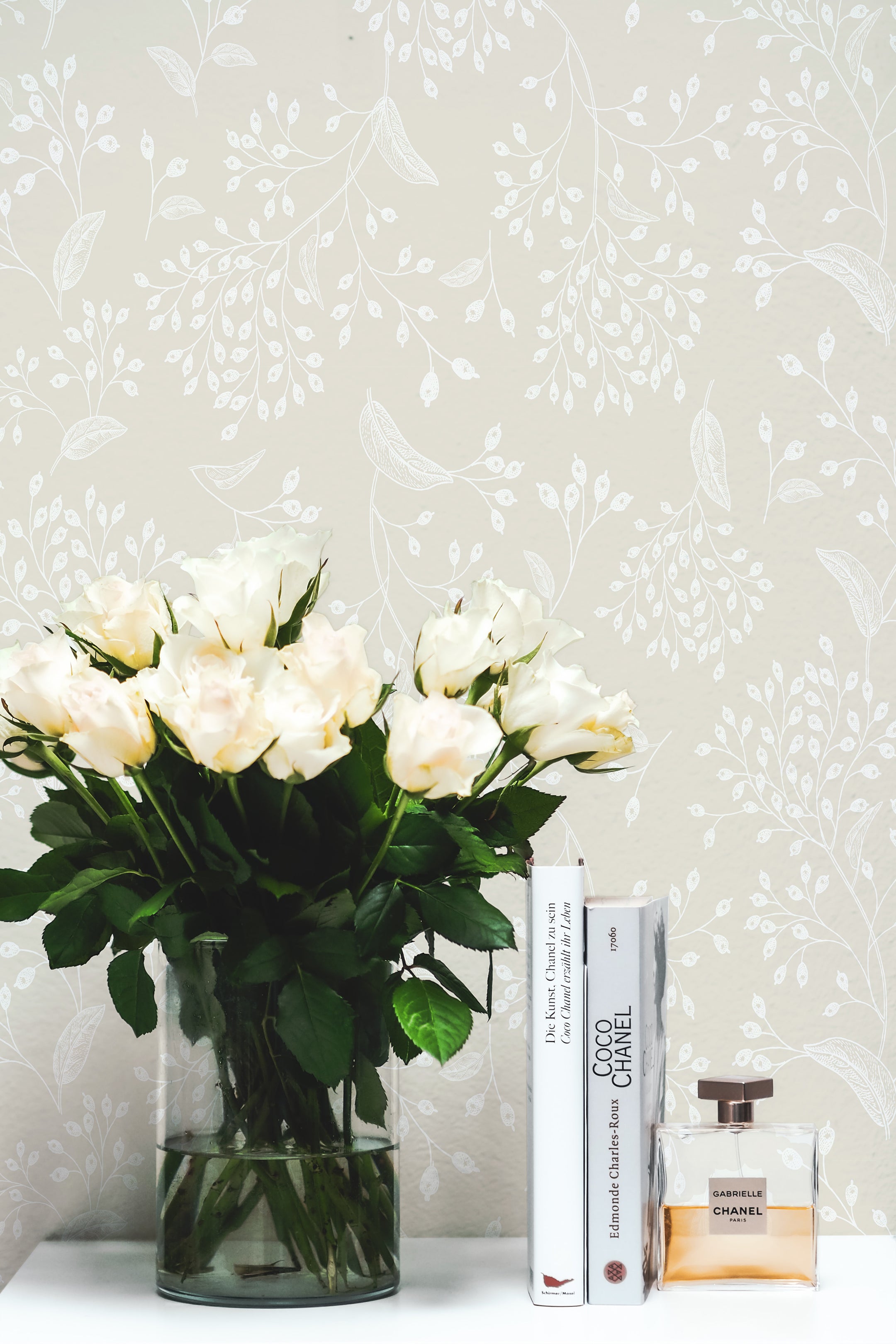 A chic and stylish room setting featuring the Botanical Whisper Wallpaper. This room includes a beige botanical patterned wallpaper, a glass vase filled with fresh white roses, and a collection of books and luxury perfume bottles, creating a refined and cultured atmosphere.
