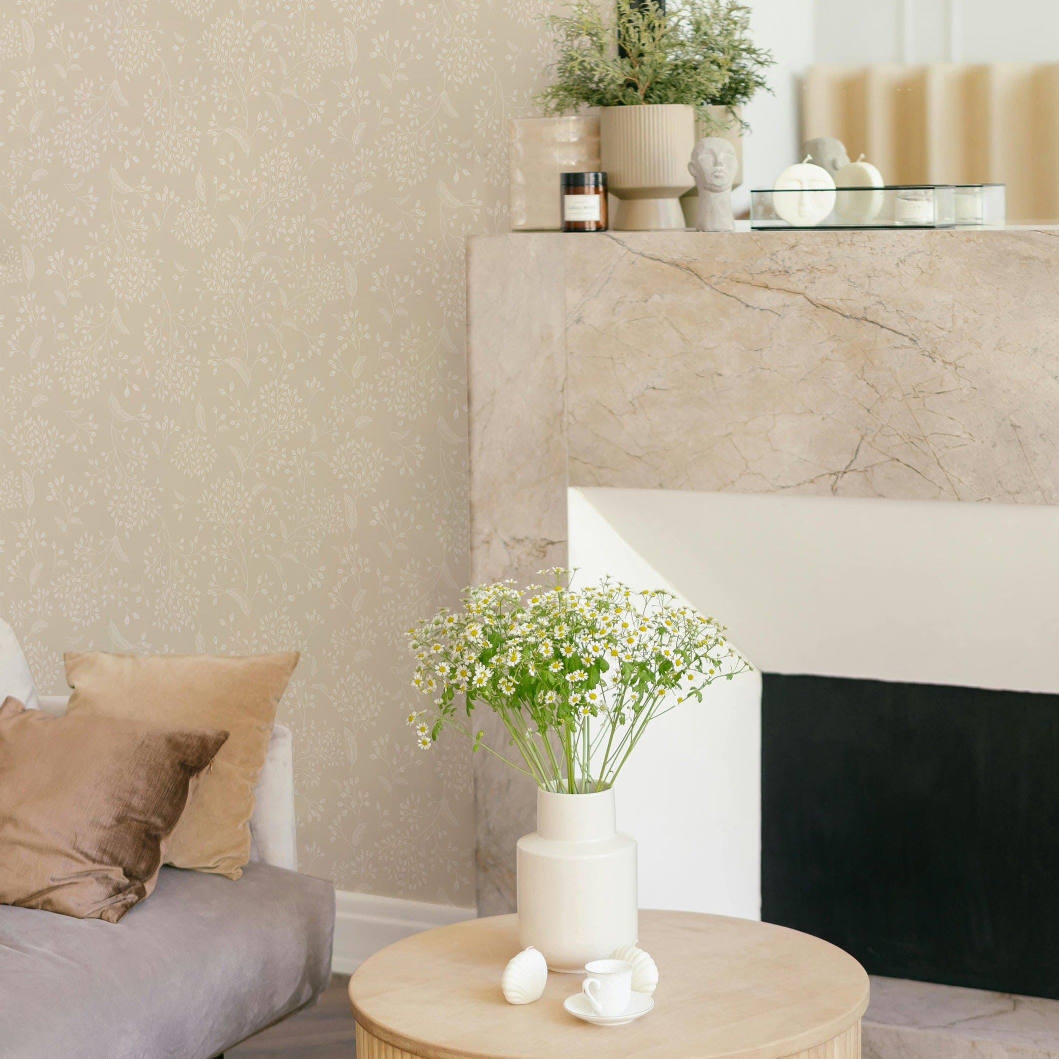 A modern living space accentuated with the Botanical Whisper Wallpaper. This wallpaper has a beige background adorned with subtle, white floral and botanical motifs, providing a soft and inviting backdrop to a contemporary room with a plush sofa, marble fireplace, and a small wooden coffee table with a vase of white flowers