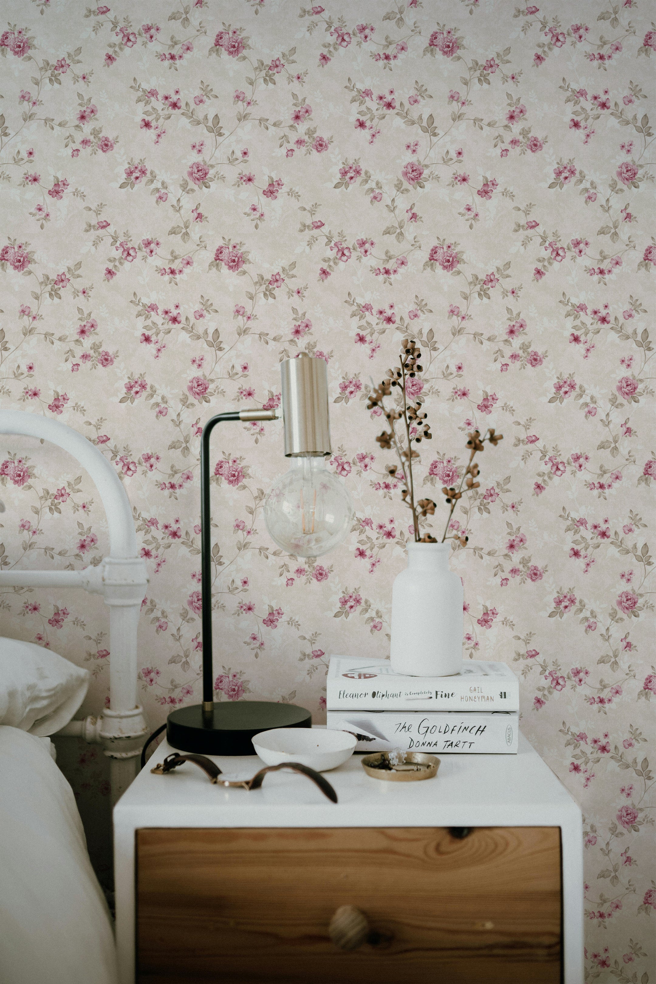 A cozy bedroom setting showcasing the Catrice Floral Wallpaper, which features a delicate pattern of pink roses and soft green foliage on a light beige background. The wallpaper lends a vintage charm to the room, complemented by a modern bedside lamp and a simple white vase with sprigs.