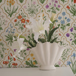 A stylish home decor scene featuring a decorative floral wallpaper with a rich array of garden flowers against a structured, geometric backdrop. The scene is accentuated with a white vase holding fresh white lilies, emphasizing the elegance and freshness of the wallpaper design.