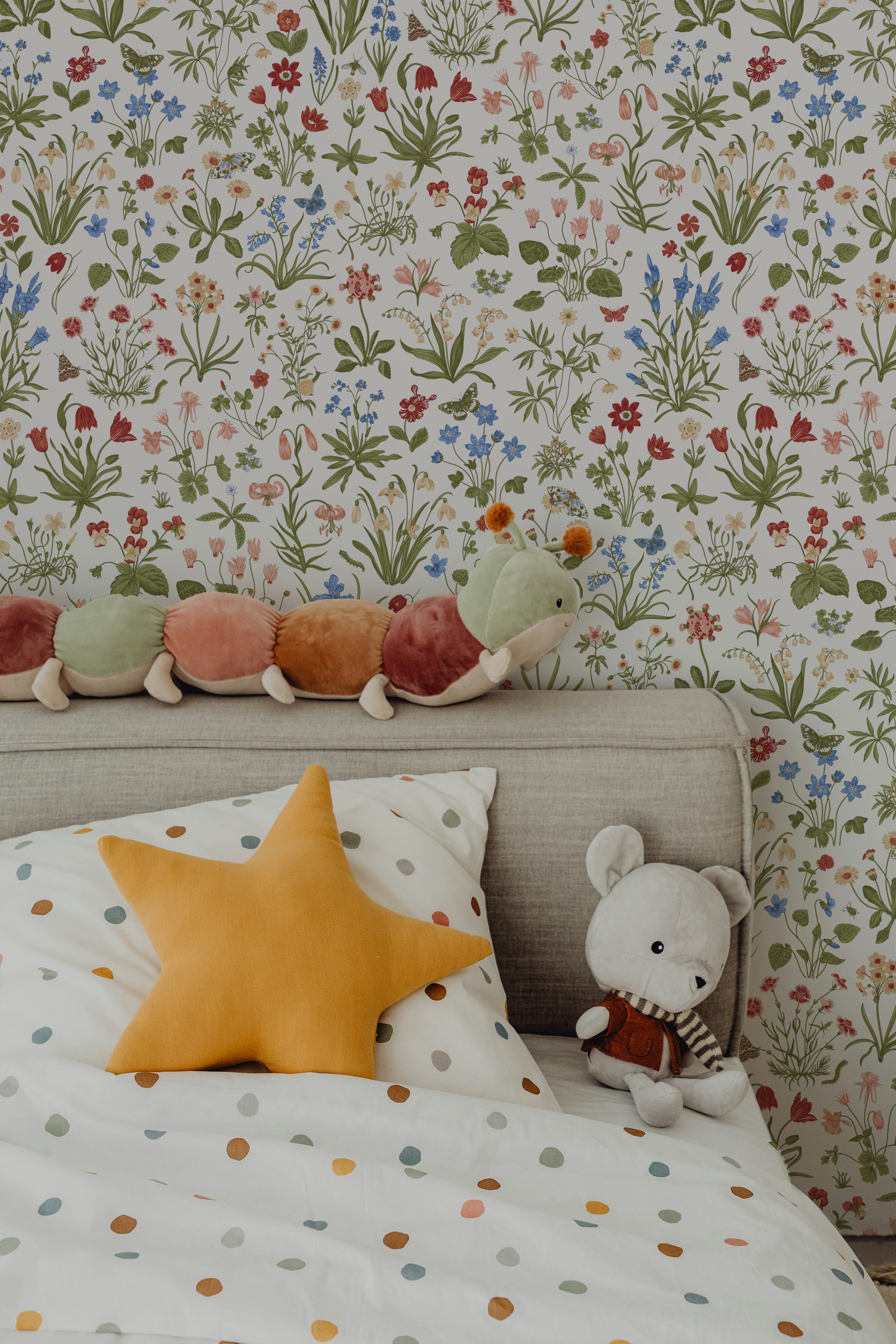 Nursery room featuring 'Garden Fantasy Wallpaper' with a diverse array of hand-drawn wildflowers in vibrant colors on a cream background, adding a joyful and colorful ambiance. A colorful caterpillar plush toy sits atop the bed, complementing the playful and nature-inspired theme of the room.