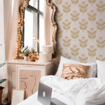A serene and stylish bedroom featuring the Ditsy Daisy Wallpaper, adorned with delicate pink daisies and golden leaves on a pale background. The room includes a vintage gold-framed mirror, white bedding, and cozy decorative elements, creating a warm and inviting atmosphere.