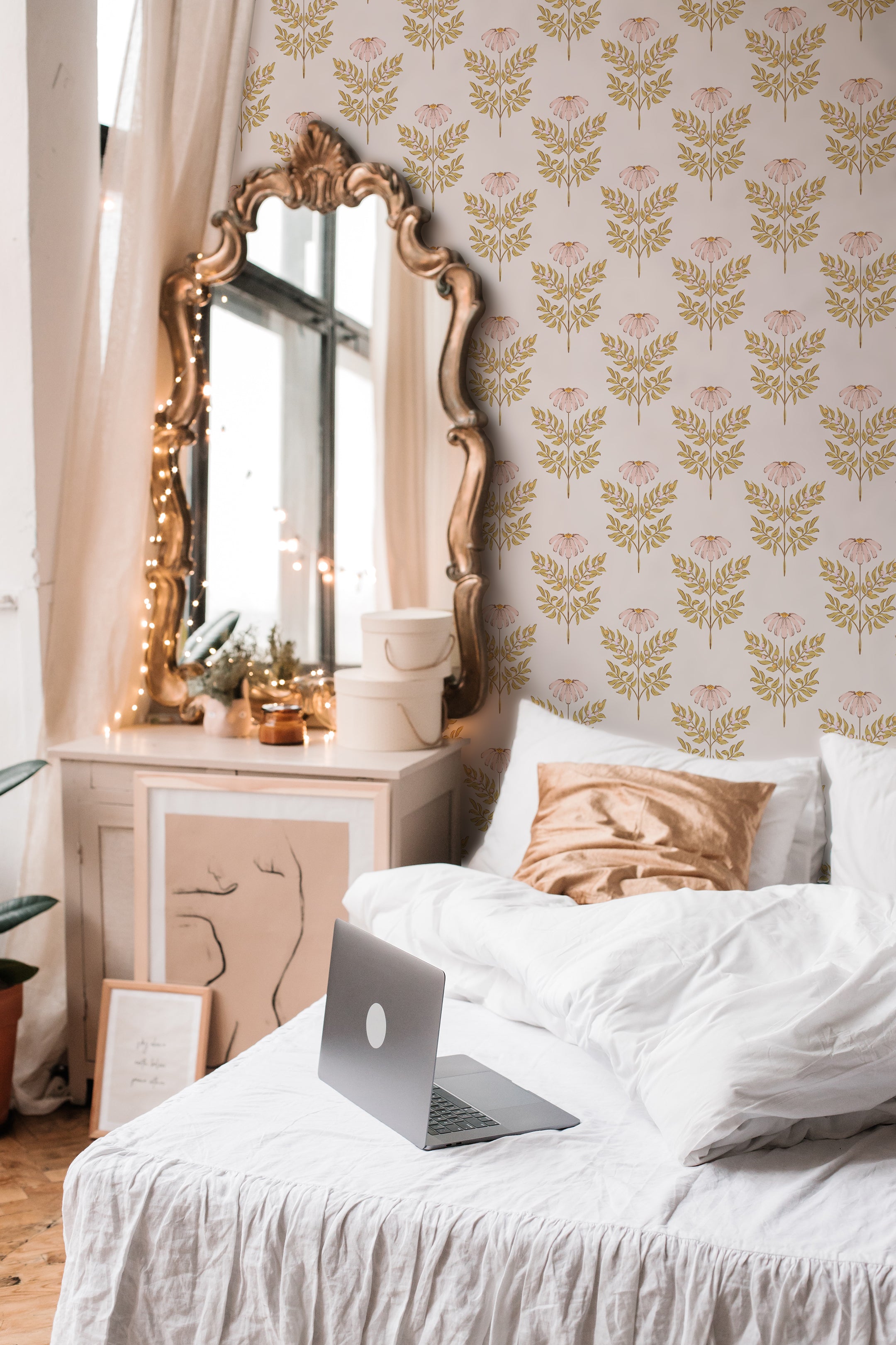A serene and stylish bedroom featuring the Ditsy Daisy Wallpaper, adorned with delicate pink daisies and golden leaves on a pale background. The room includes a vintage gold-framed mirror, white bedding, and cozy decorative elements, creating a warm and inviting atmosphere.