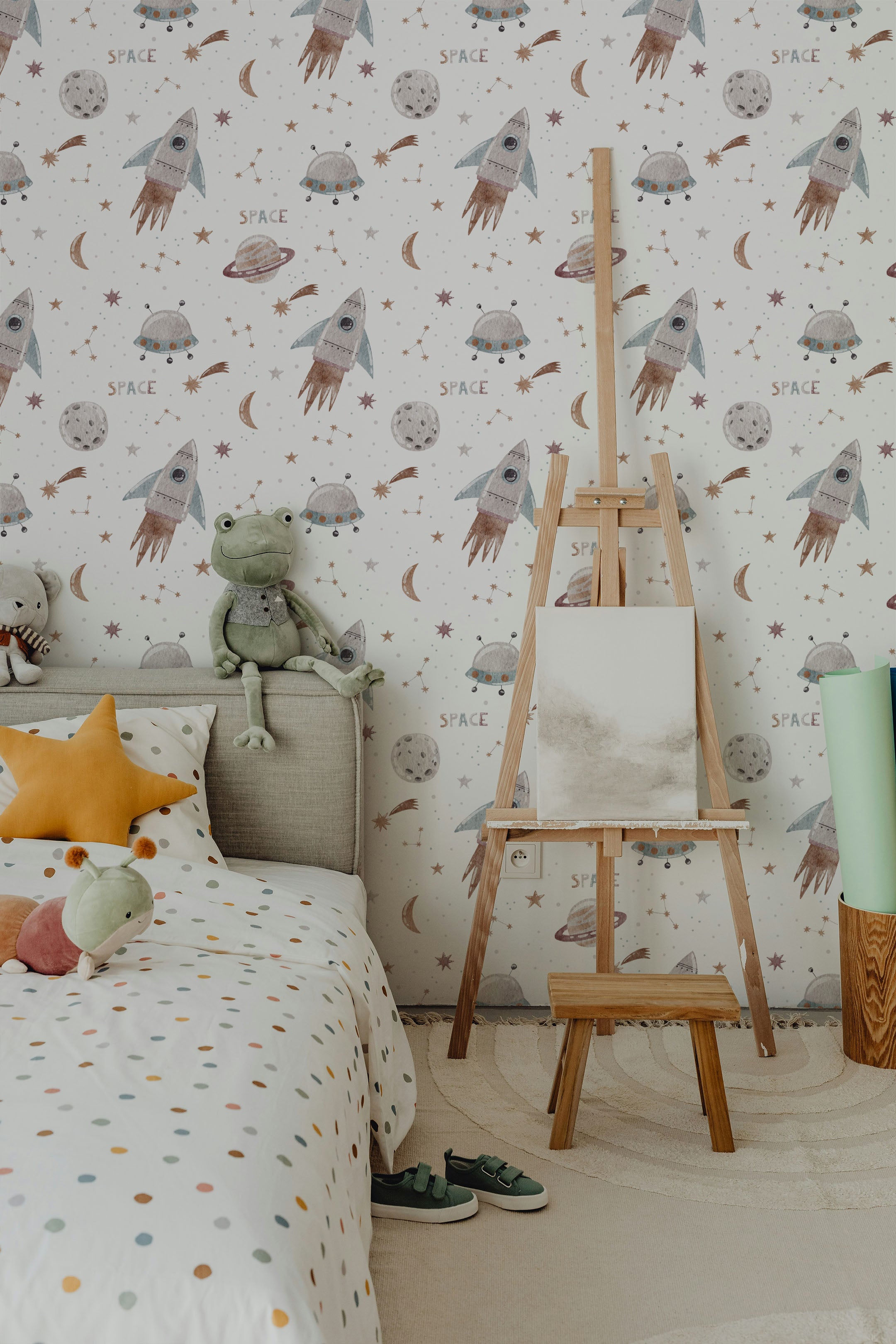 A cozy child's bedroom adorned with Space Craze Wallpaper, featuring cute space motifs like rockets, satellites, and planets in a gentle color palette. The room is styled with a bed, colorful pillows, and a wooden easel, creating an inviting and imaginative play area.