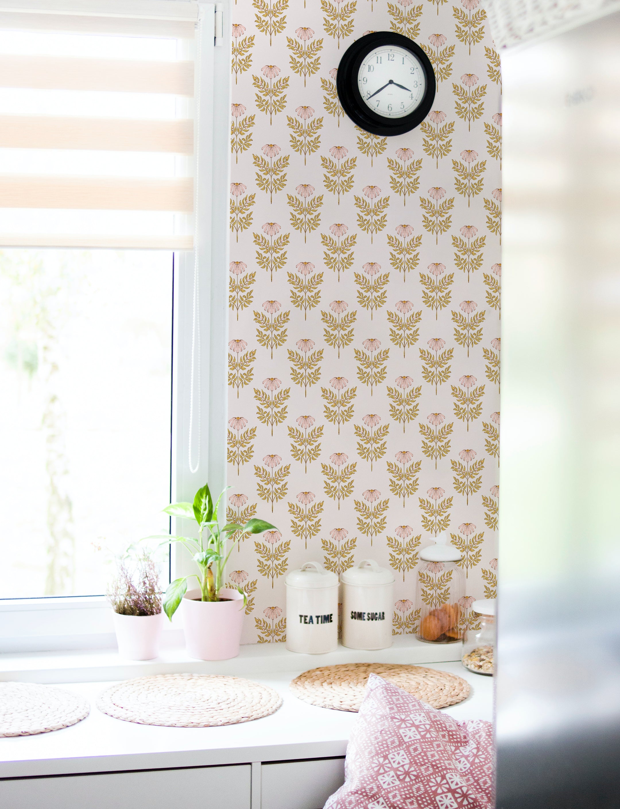 A cozy kitchen nook enhanced by the Ditsy Daisy Wallpaper - Small, featuring elegant small pink flowers and golden leaves on a soft background. The area is styled with a modern white bench, woven placemats, and simple kitchen canisters, creating a warm and inviting space.