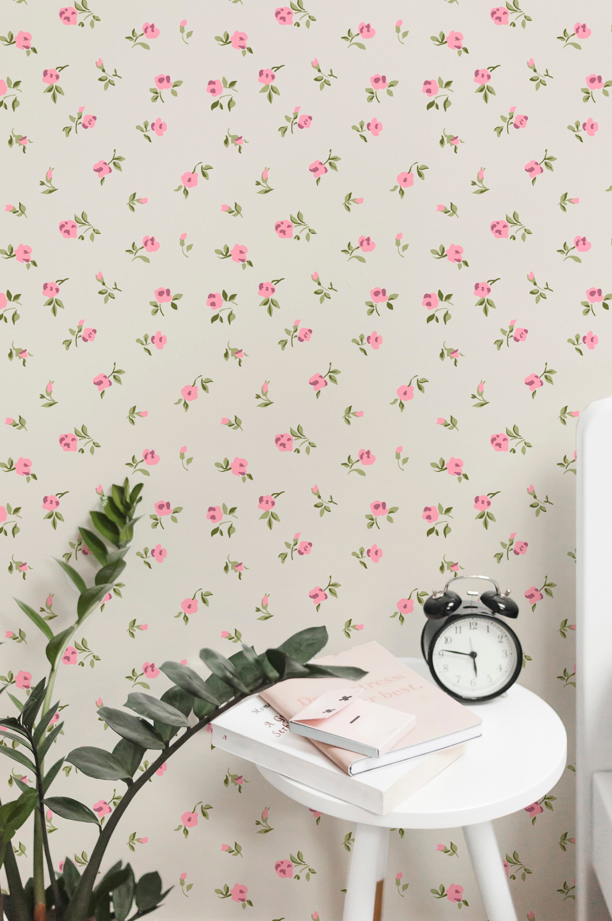 A refreshing and lively workspace featuring the Flora Wallpaper, adorned with small, vibrant pink blossoms and lush green leaves on a pale cream background. The setting includes a white side table with a stack of books and a vintage-style black alarm clock, complemented by indoor green plants for a touch of nature.