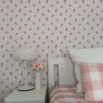 A modern bedroom adorned with Daphne Floral Wallpaper, which consists of subtle pink flowers on a light background. The room features a minimalist bedside setup with a white lamp and pink floral decorations, enhancing the soft, tranquil decor.
