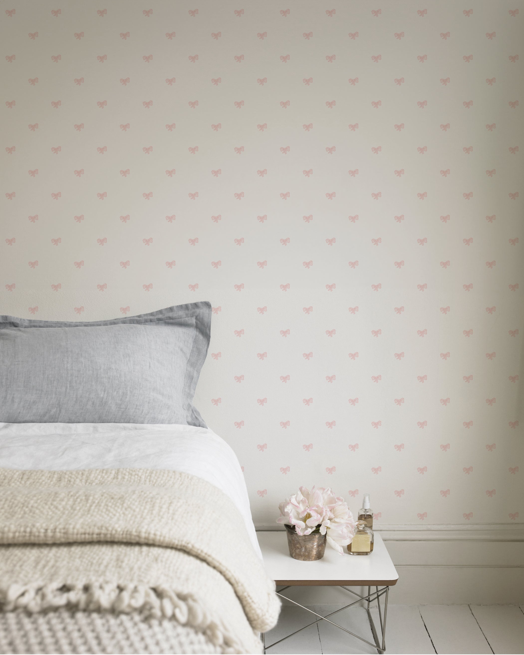 A serene bedroom decorated with the Tiny Bows Wallpaper, which has small pink bows on a light background. The decor includes a cozy bed with a gray linen, a small bedside table with a floral arrangement, and a soft knitted throw, creating a gentle and inviting atmosphere.