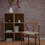 A cozy reading nook with Dusty Rose Floral Wallpaper adorning the wall, complemented by a simple wooden chair and a shelving unit filled with books. The soft pink background and white floral patterns evoke a serene, inviting space