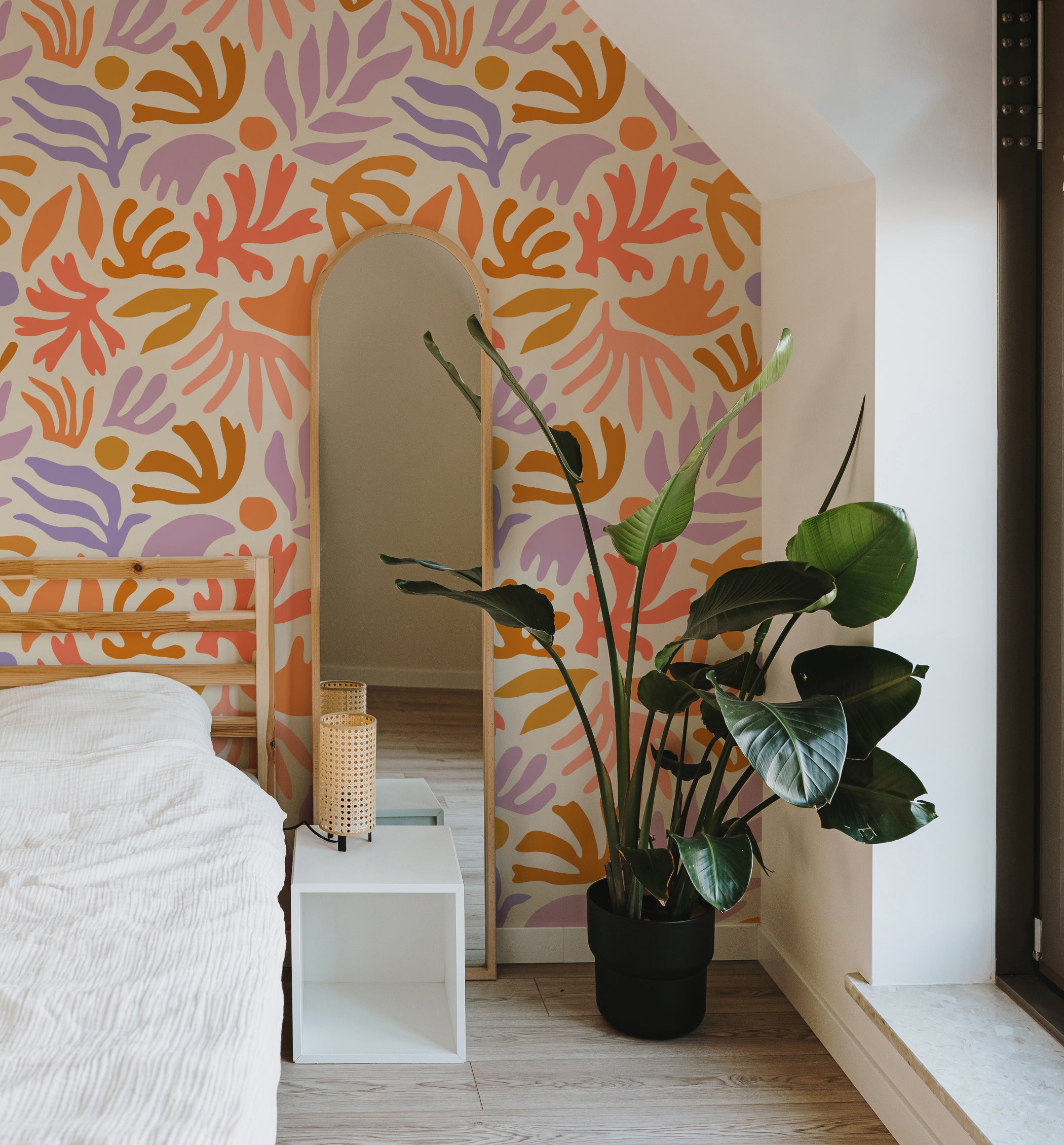 Cozy room corner showcasing an arch-shaped mirror and a large potted plant in front of a lively wallpaper with an abstract plant motif in warm tones
