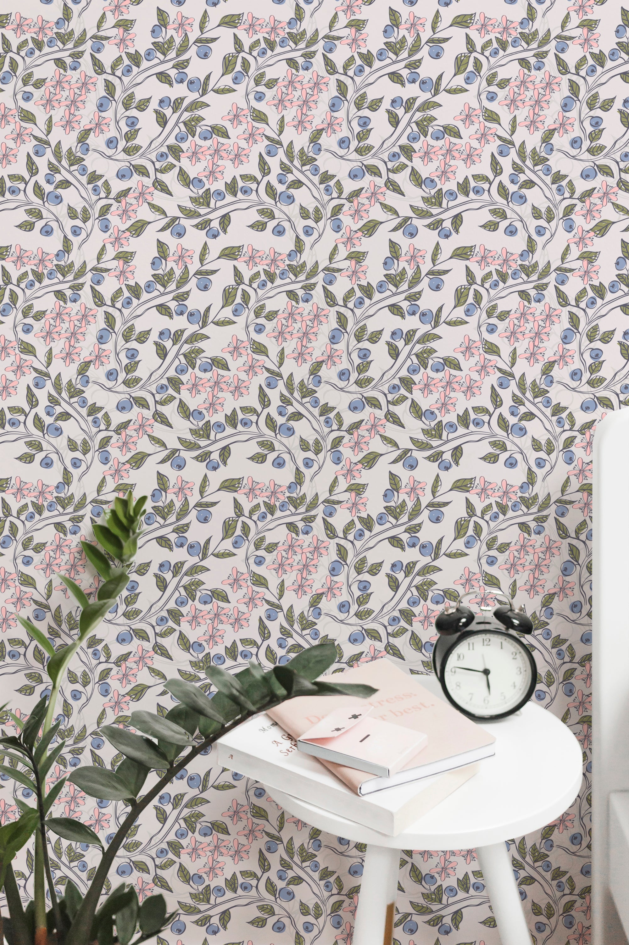 A modern workspace with a white table featuring books and a vintage clock, complemented by a wall covered in elegant wallpaper with a pattern of pink flowers and blueberries on twisting vines