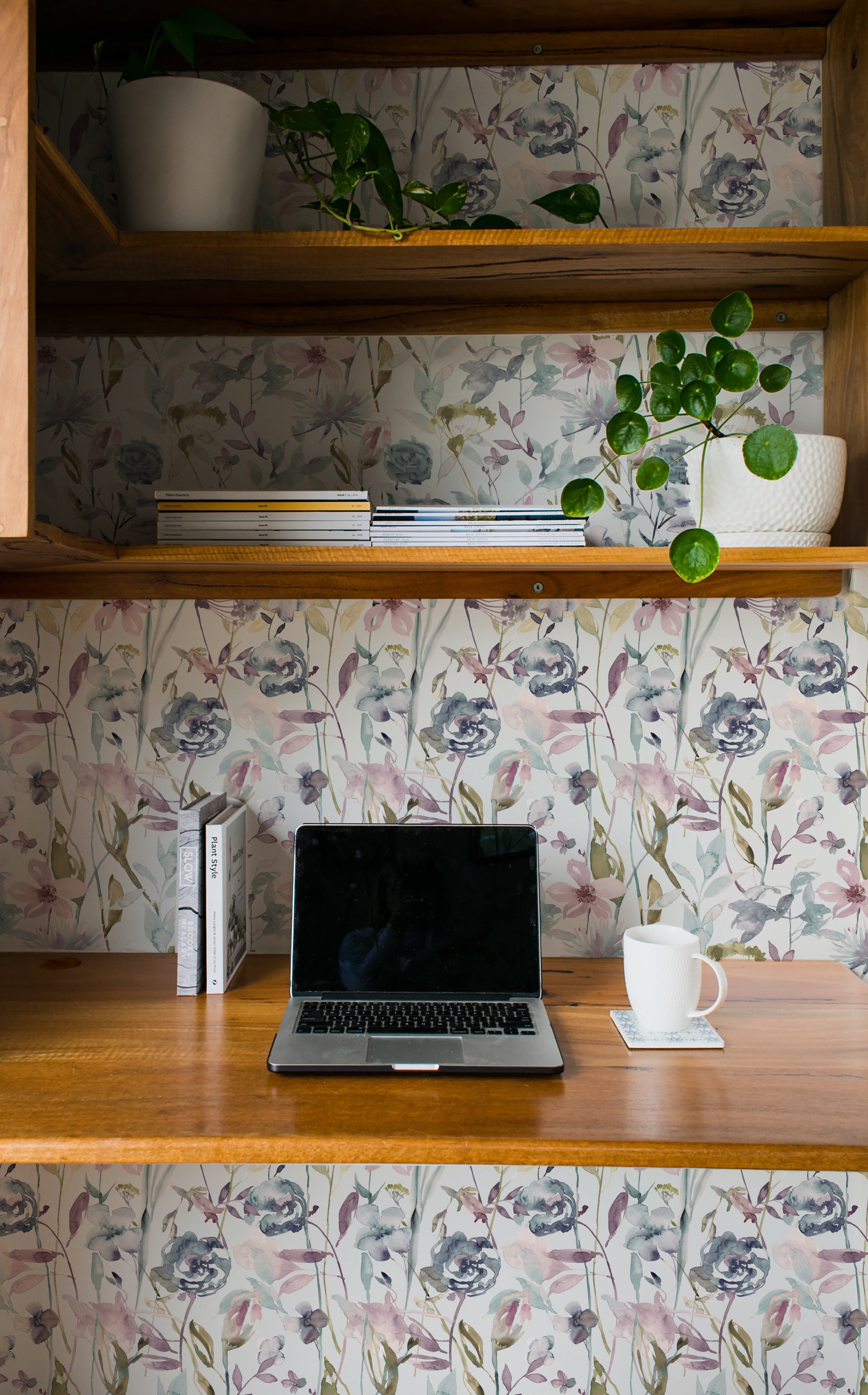 An organized home office space with a wooden desk and shelves, decorated with indoor plants and books. The wall behind the desk is covered in Watercolour Meadow Wallpaper, displaying delicate flowers in muted watercolor hues, creating a tranquil and inspiring work environment.