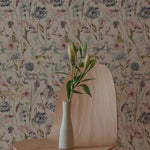 A minimalistic interior featuring a modern chair with a vase of fresh lilies on it. The background showcases the Watercolour Meadow Wallpaper, with its soft watercolor floral pattern in shades of pink, blue, and green, giving a serene and artistic touch to the space.
