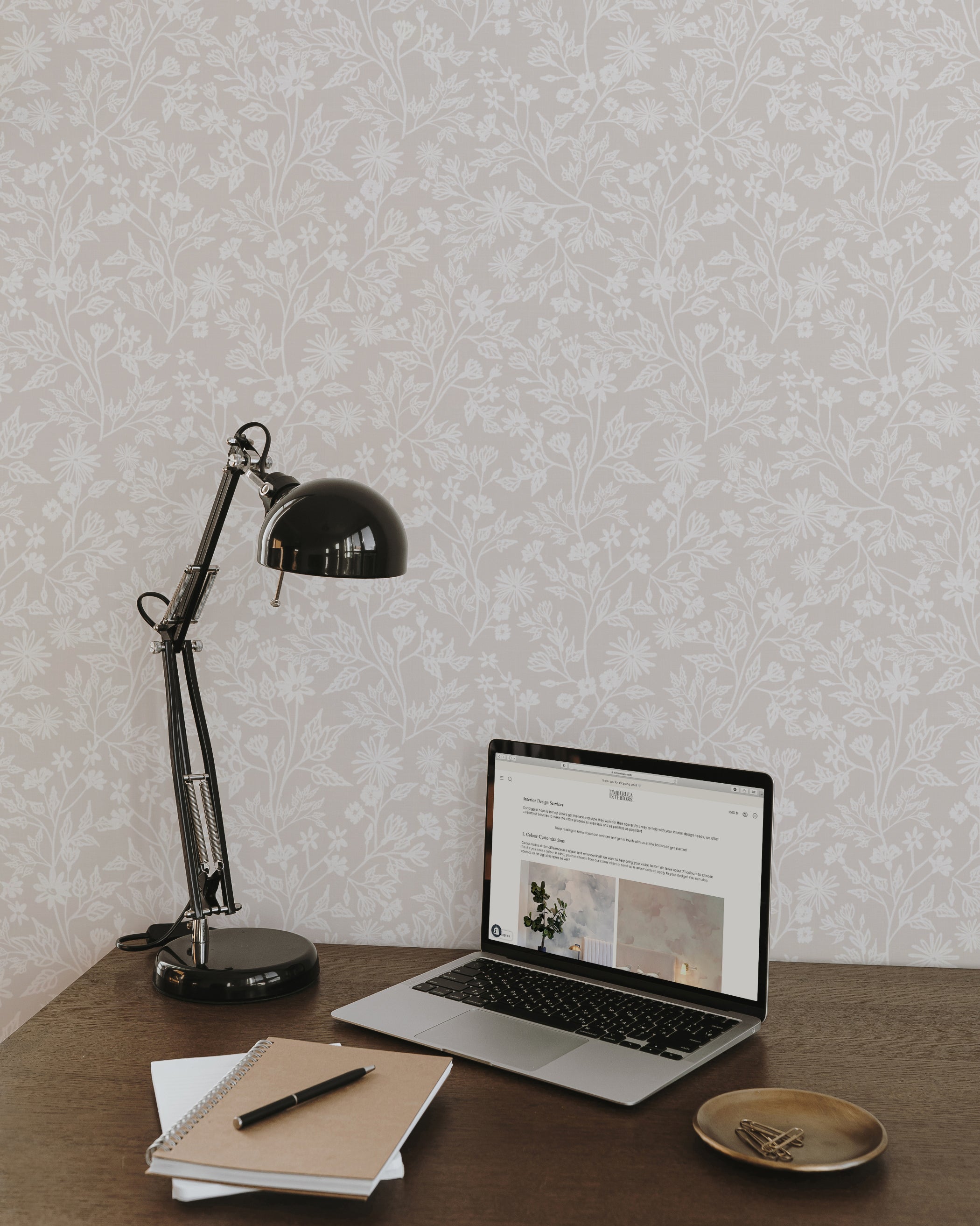 A stylish home office featuring Blossom Haze Wallpaper, decorated with a subtle floral pattern in shades of light gray on a white background. The elegant wallpaper enhances the modern workspace equipped with a black desk lamp, laptop, and notebooks.