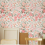 A creative space with a table covered in paper cutouts and crafts supplies, surrounded by chairs. The background wall is adorned with the Floral Frenzy Wallpaper, which has a lively pattern of pink, peach, and beige flowers amidst green leaves.