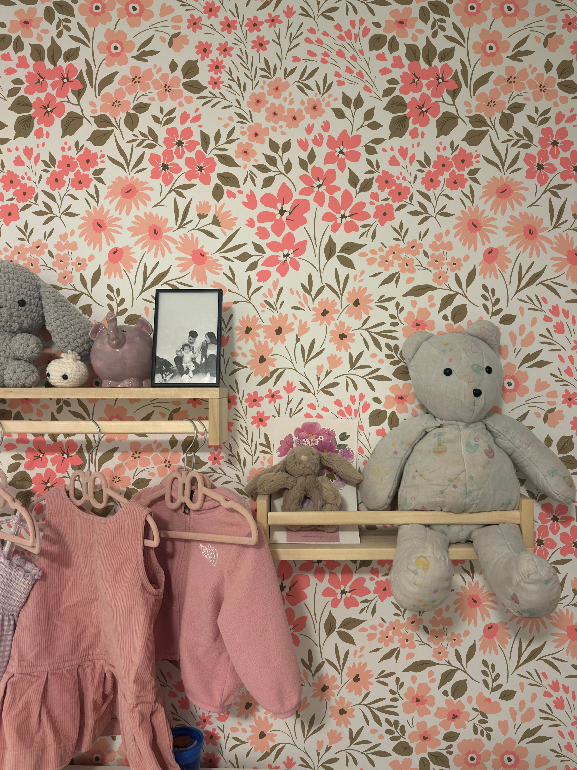 A cozy corner of a child's room with a shelf displaying various plush toys, a framed photo, and children's clothing hanging below. The wall is covered in Floral Frenzy Wallpaper, featuring a dense pattern of pink and peach flowers with green and brown foliage.