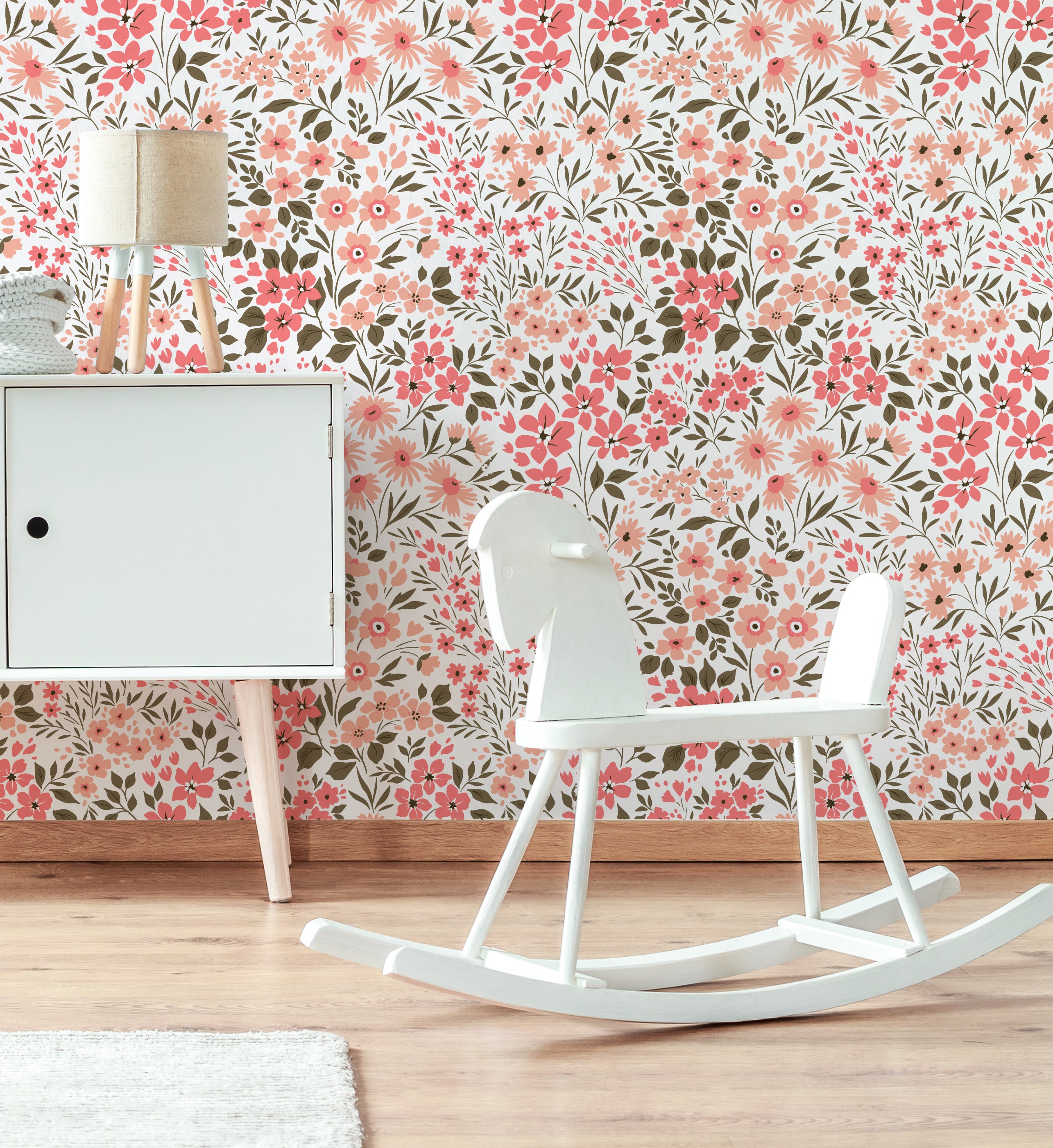 A nursery room featuring a white rocking horse in front of a small cabinet with a lamp on top, beside a plush rug. The room is brightened by the Floral Frenzy Wallpaper, showing a vibrant display of pink and peach flowers against a backdrop of green leaves.