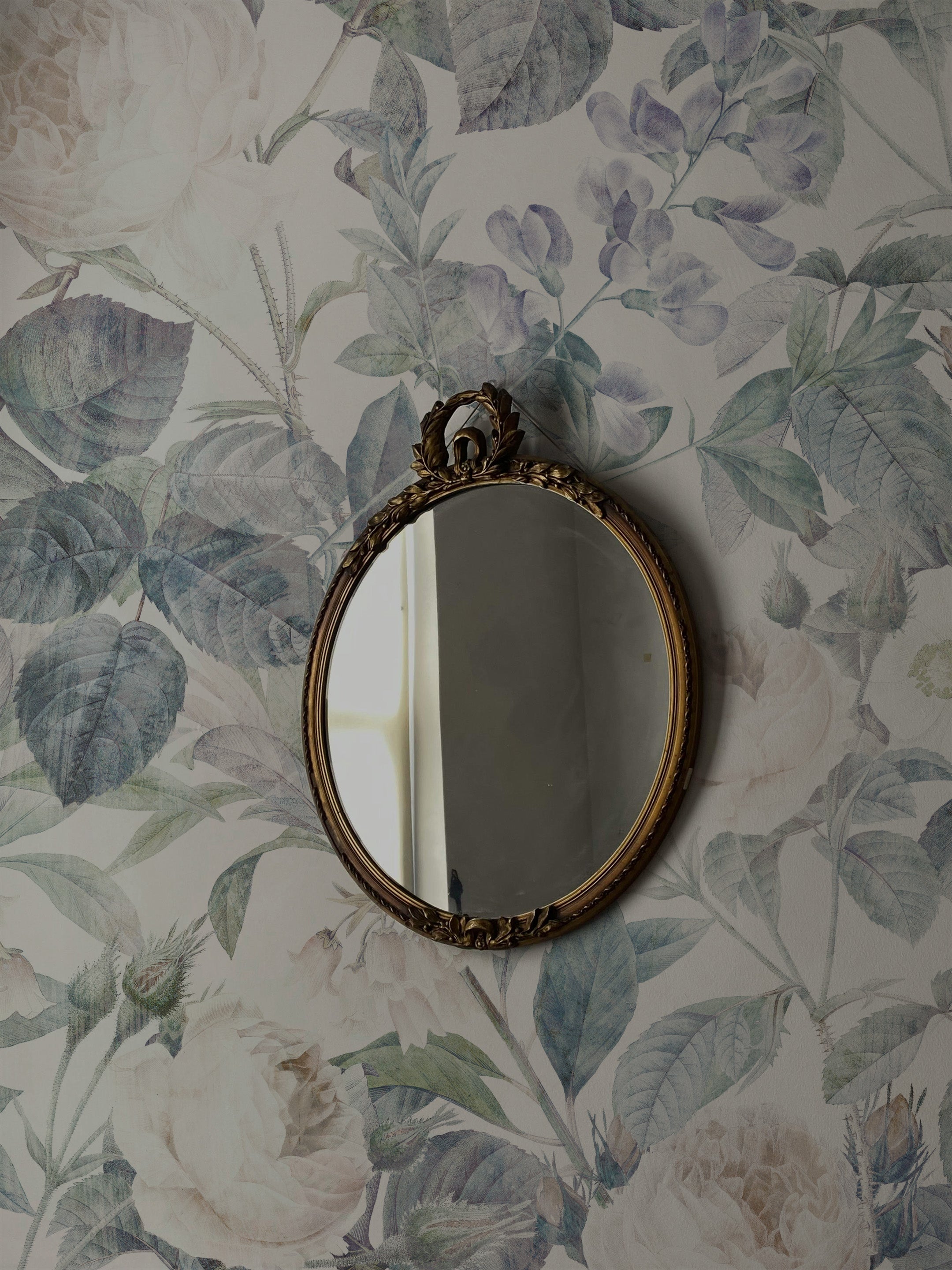 Detail of the Faded Luxury Wallpaper with a vintage ornate mirror reflecting its large, soft-hued floral patterns. The wallpaper's faded effect on the flowers and leaves provides a timeless elegance, contributing to a sophisticated and peaceful room decor.